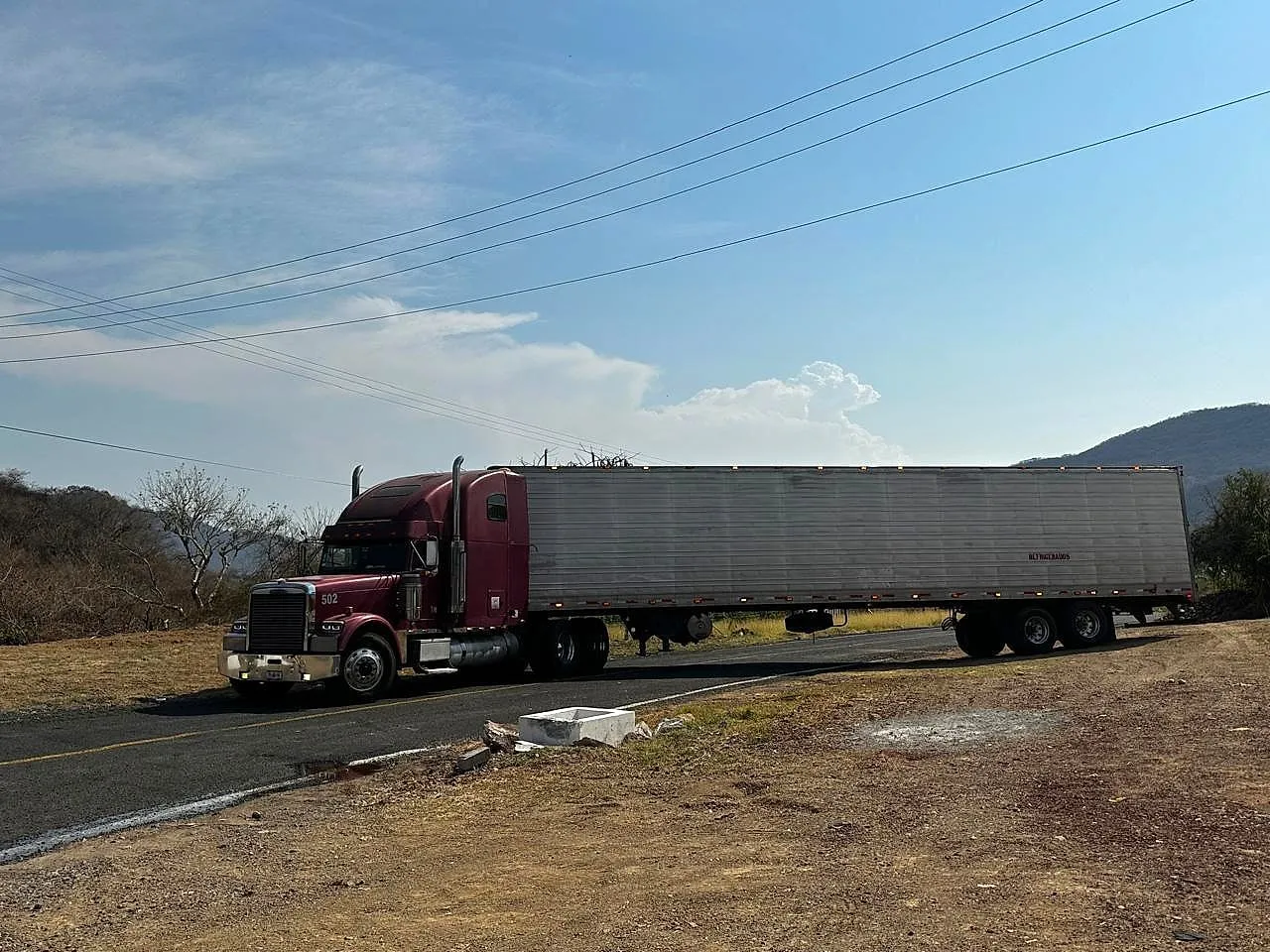 Tractocamión guinda número 502 con caja refrigerada, detenido en una carretera entre cerros.